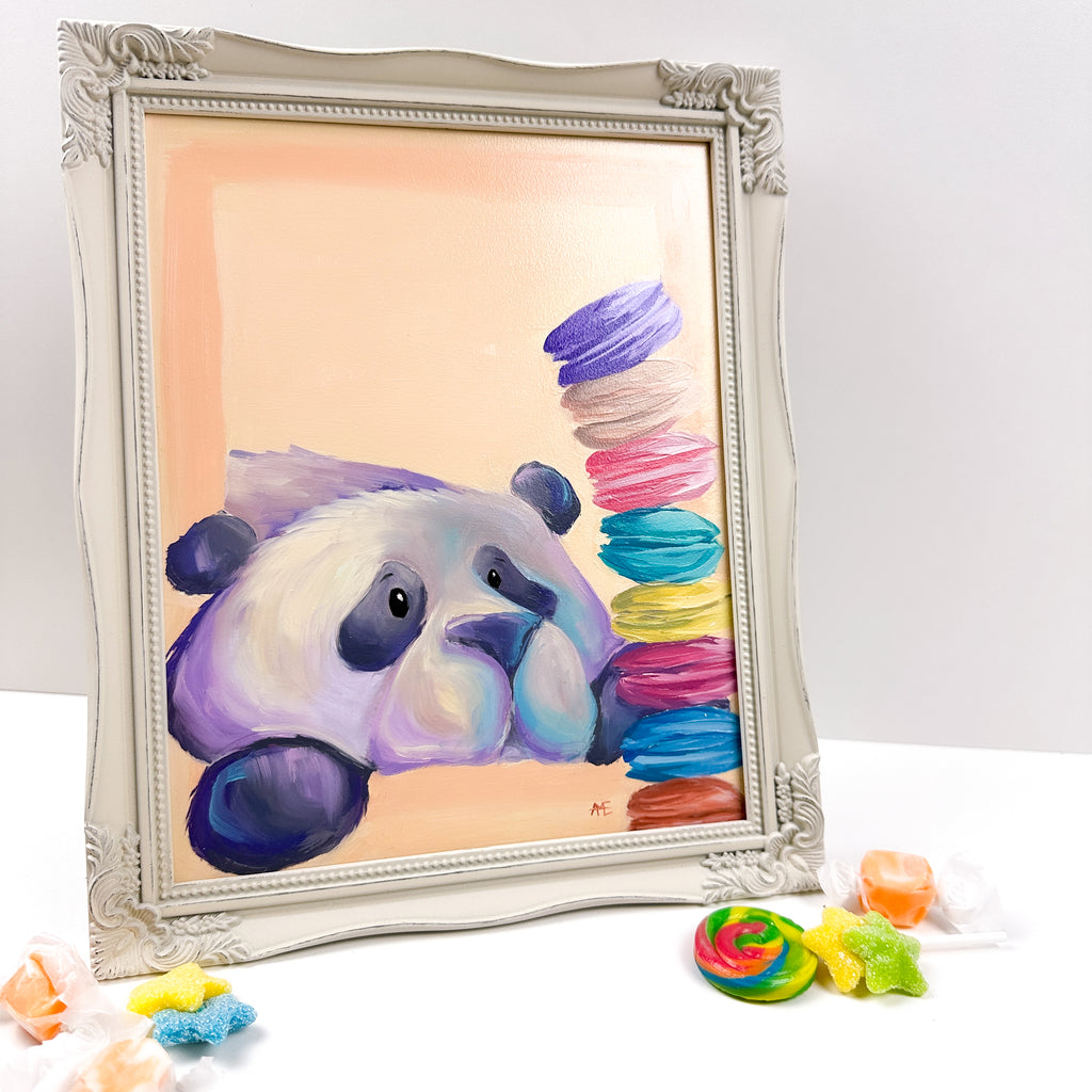 framed painting with a peach colored background of a cute kawaii/illustrative style panda, staring at a stack of colorful macarons.