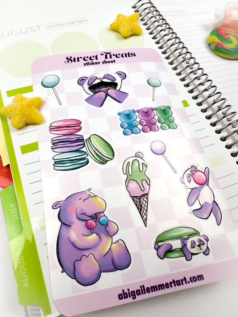 A sticker sheet with 3 cake pops, a stack of macaron, the panda with mouthful of macarons, hippo with cake pops, ice cream, panda macaron sandwich , dancing bubble gum panda and gummy bears sitting on top of an open daily planner.
