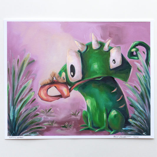 A colorful art print featuring a green cartoon lizard character with large white eyes and a long pink tongue sticking out, surrounded by stylized green plants on a pink-purple background. The print is shown against a clean white backdrop.