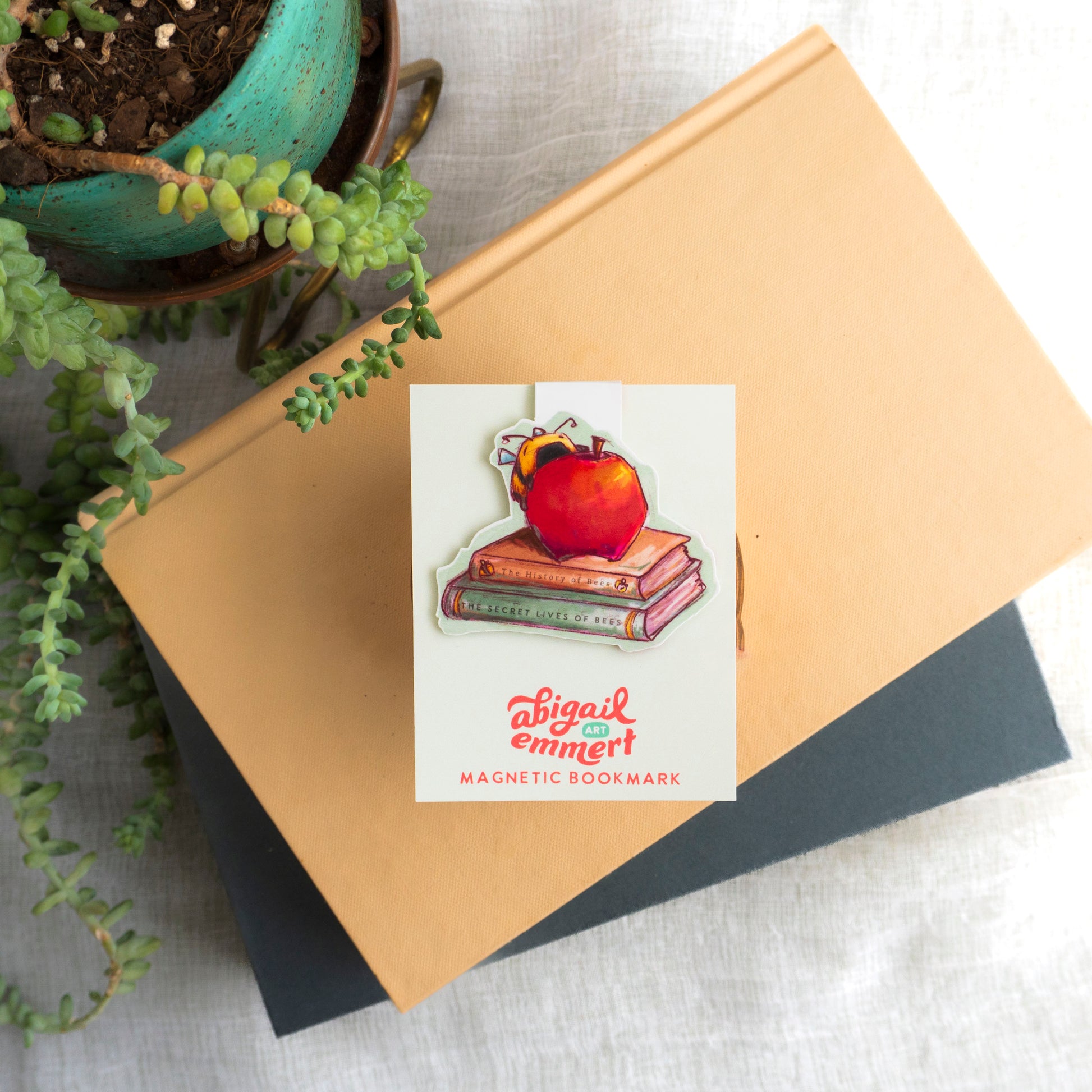 Magnetic bookmark with a design of an apple on books, placed on a notebook with a potted plant in the background.