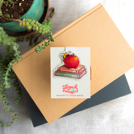 Magnetic bookmark with a design of an apple on books, placed on a notebook with a potted plant in the background.