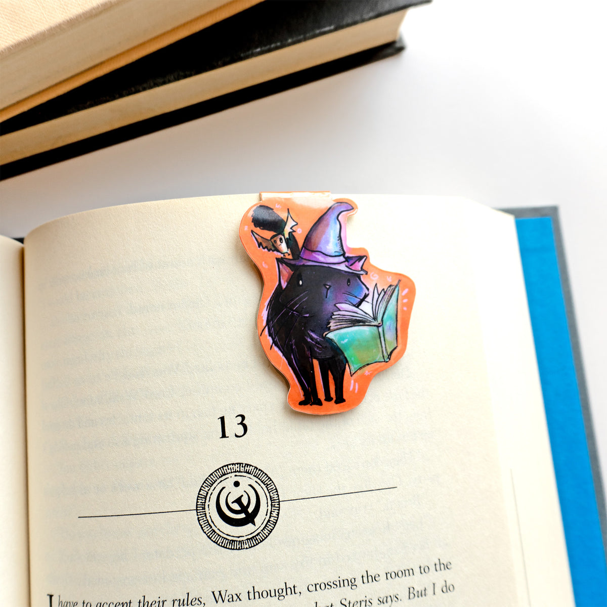 magnetic bookmark shown holding a space in a book of an image of a black cat wearing a purple witches hat with a bat overhead and spellbook hovering in front of it. 