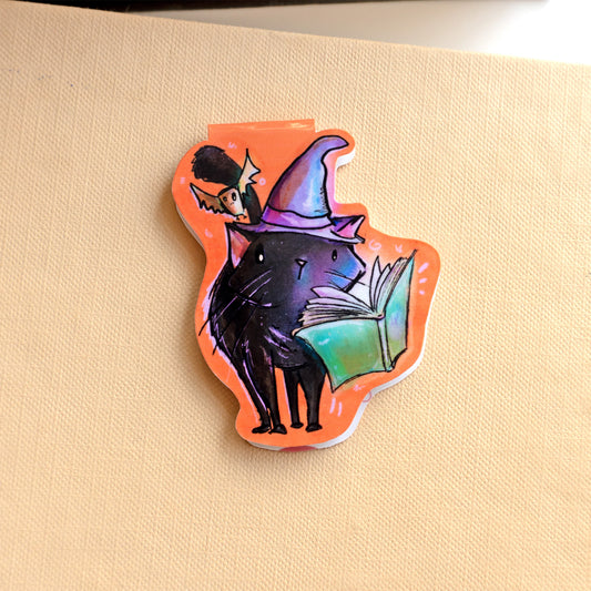 magnetic bookmark image of a black cat wearing a purple witches hat with a bat overhead and spellbook hovering in front of it. 