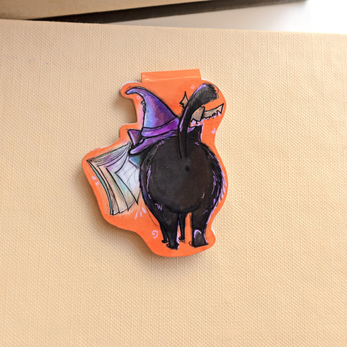 back of magnetic bookmark image of a black cat wearing a purple witches hat with a bat overhead and spellbook hovering in front of it. 