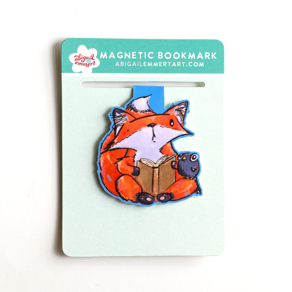 A magnetic bookmark by Abigail Emmert Art featuring a cartoon fox reading a book with a small crow friend, packaged on a mint green backing card.