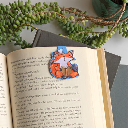 A small orange fox bookmark with a book and a small crow friend, placed on an open book page. The book is styled with a gray folder and green trailing leaves in the background.
