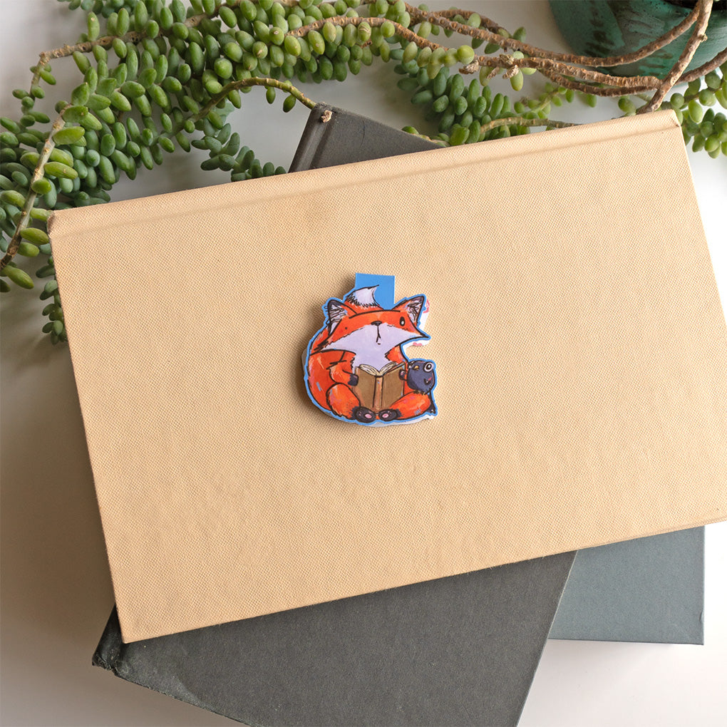 A small orange fox bookmark with a book and a small crow friend. Set against a kraft paper notebook on a dark gray book with green trailing succulents in the background.