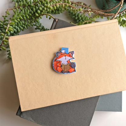 A small orange fox bookmark with a book and a small crow friend. Set against a kraft paper notebook on a dark gray book with green trailing succulents in the background.