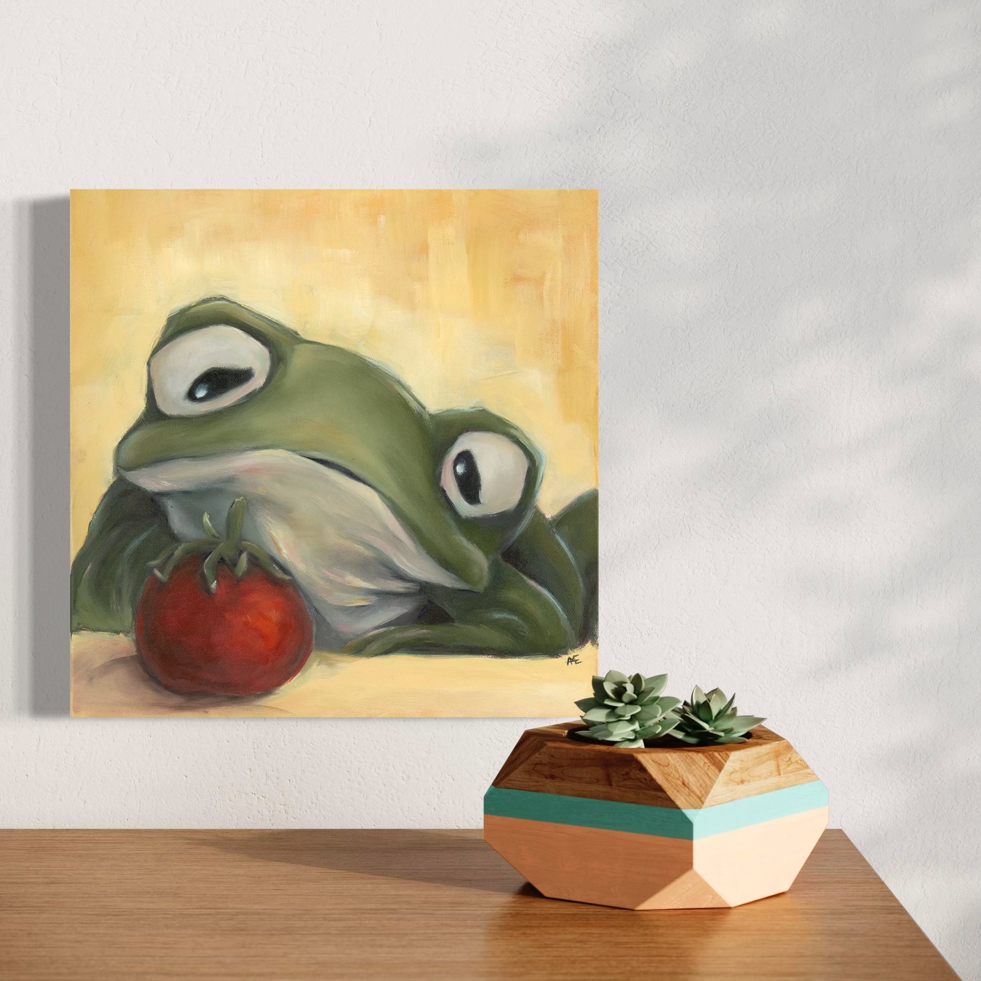 painting of a frog staring longingly at a cherry tomato hanging on a wall above a wooden surface with a geometric plant holder.
