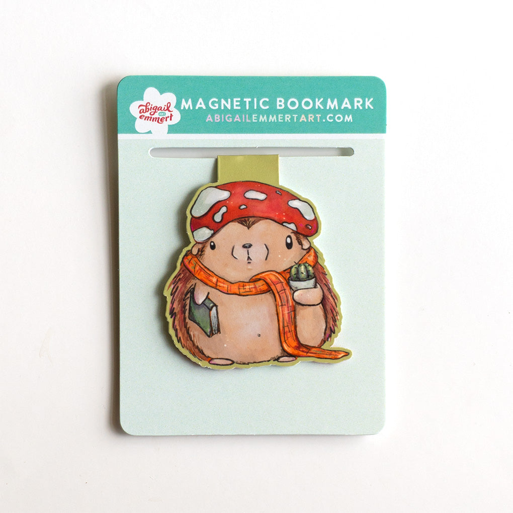 A cute whimsical hedgehog magnetic bookmark by Abigail Emmert Art, featuring a red mushroom cap, orange scarf, and green book, with his cactus friend. The bookmark is packaged on a light green backing card.