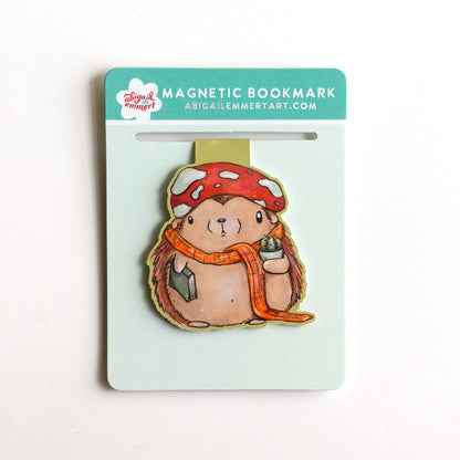 A cute whimsical hedgehog magnetic bookmark by Abigail Emmert Art, featuring a red mushroom cap, orange scarf, and green book, with his cactus friend. The bookmark is packaged on a light green backing card.
