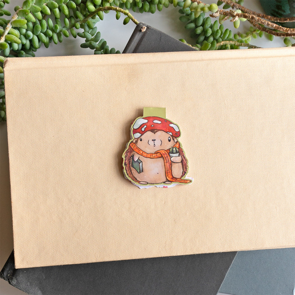 A cute whimsical hedgehog magnetic bookmark by Abigail Emmert Art, featuring a red mushroom cap, orange scarf, and green book, with his cactus friend. placed on top of a beige textured book with green trailing leaves around it.