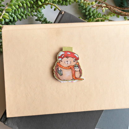 A cute whimsical hedgehog magnetic bookmark by Abigail Emmert Art, featuring a red mushroom cap, orange scarf, and green book, with his cactus friend. placed on top of a beige textured book with green trailing leaves around it.