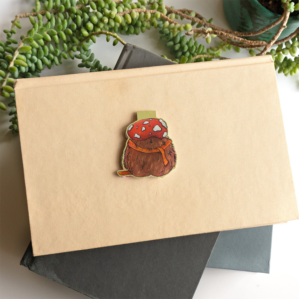 The back side of a cute whimsical hedgehog magnetic bookmark by Abigail Emmert Art, featuring a red mushroom cap, orange scarf, and green book, with his cactus friend.placed on top of a beige book with dark gray and green books underneath and trailing green leaves around the edges.