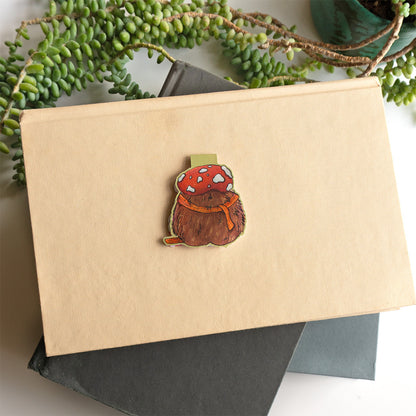 The back side of a cute whimsical hedgehog magnetic bookmark by Abigail Emmert Art, featuring a red mushroom cap, orange scarf, and green book, with his cactus friend.placed on top of a beige book with dark gray and green books underneath and trailing green leaves around the edges.