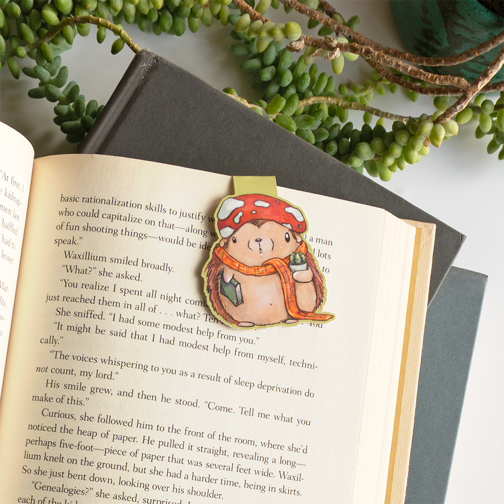 A cute whimsical hedgehog magnetic bookmark by Abigail Emmert Art, featuring a red mushroom cap, orange scarf, and green book, with his cactus friend.