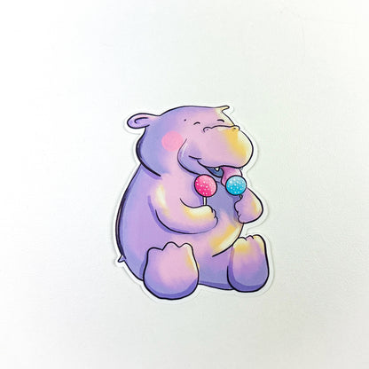 An illustrated sticker of a happy purple hippo licking a blue cake pop and holding a pink one in it's other paw.