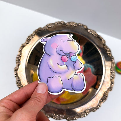 An illustrated sticker of a happy purple hippo licking a blue cake pop and holding a pink one in it's other paw.