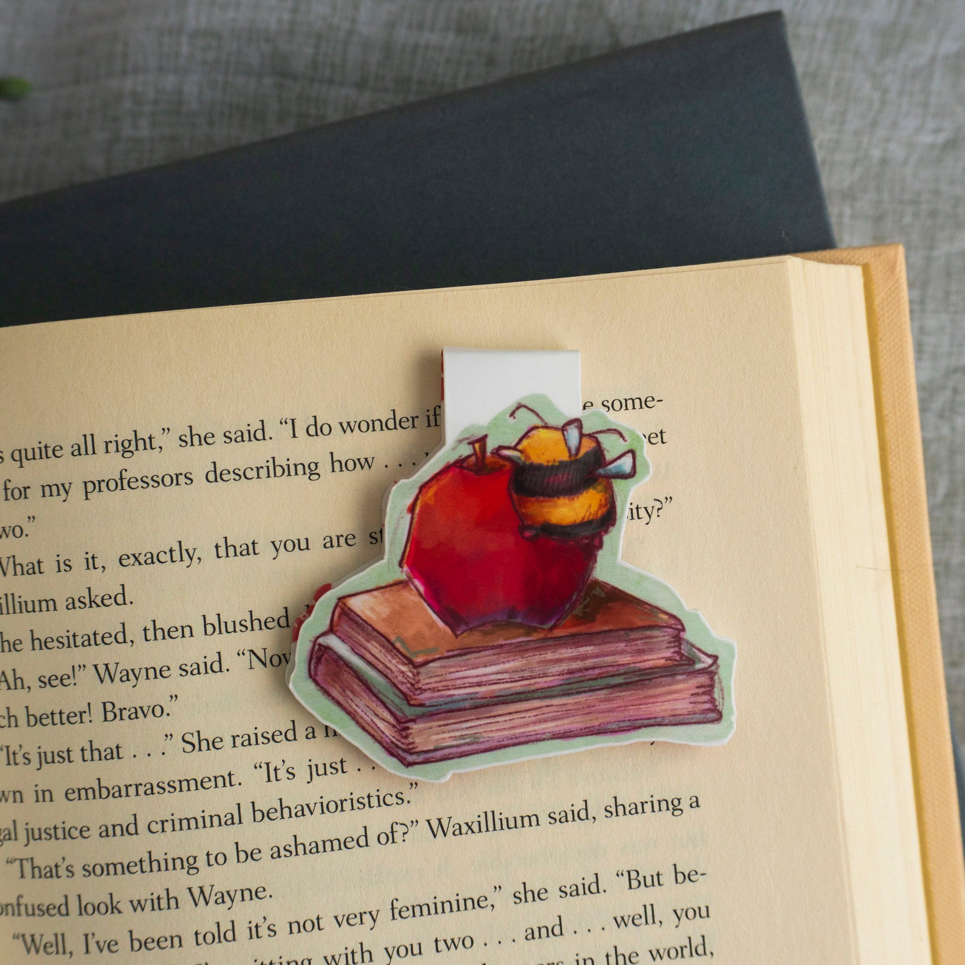 Open book with a sticker of a bee on a book and apple, placed on a textured surface.