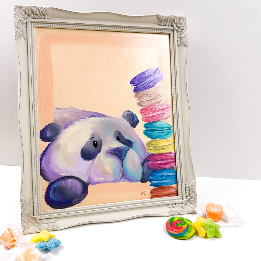 framed painting with a peach colored background of a cute kawaii/illustrative style panda, staring at a stack of colorful macarons. 