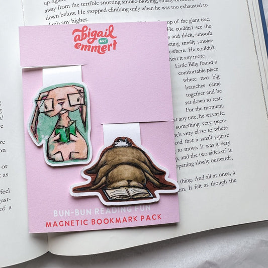 two bunnies as bookmarks, One peach/pink colored bunny wearing glasses and reading a book. And a brown droopy bunny laying on its belly with it's face smashed in a book. 