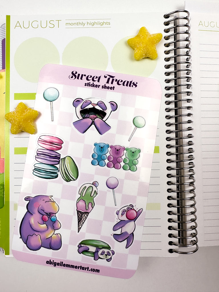 A sticker sheet with 3 cake pops, a stack of macaron, the panda with mouthful of macarons, hippo with cake pops, ice cream, panda macaron sandwich , dancing bubble gum panda and gummy bears sitting on top of an open daily planner