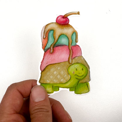 An illustrated sticker of a green turtle with a waffle cone shell and pink, blue and orange ice cream melting on top along with Carmel sauce dripping and a cherry on top!