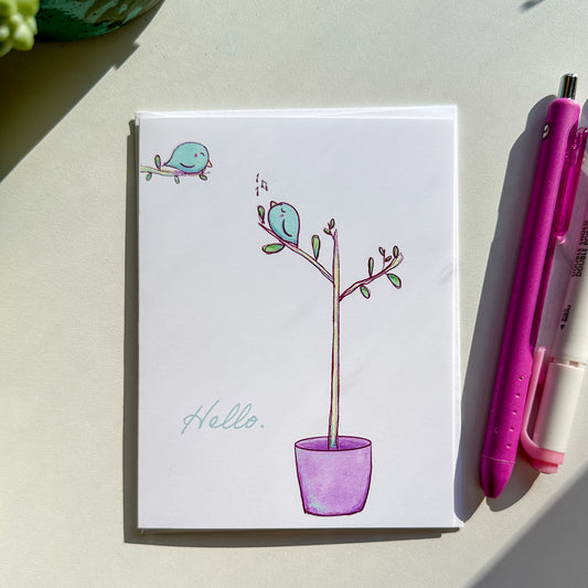 Greeting card in an illustrative style of a blue bird in the upper left corner on a branch and another bird on a potted sapling with music notes near it.  The word HELLO. is written in the bottom left in a script font and light blue. The greeting card is pictured  next to a pen.