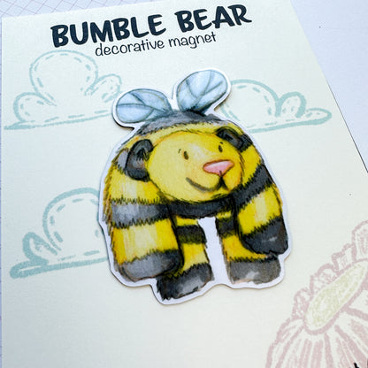 magnet of a yellow and black bear with wings that is flying in the sky but the body is too heavy so he droops a bit.  Magnet is pictured on the backing card which has an illustration of clouds and flowers behind it.