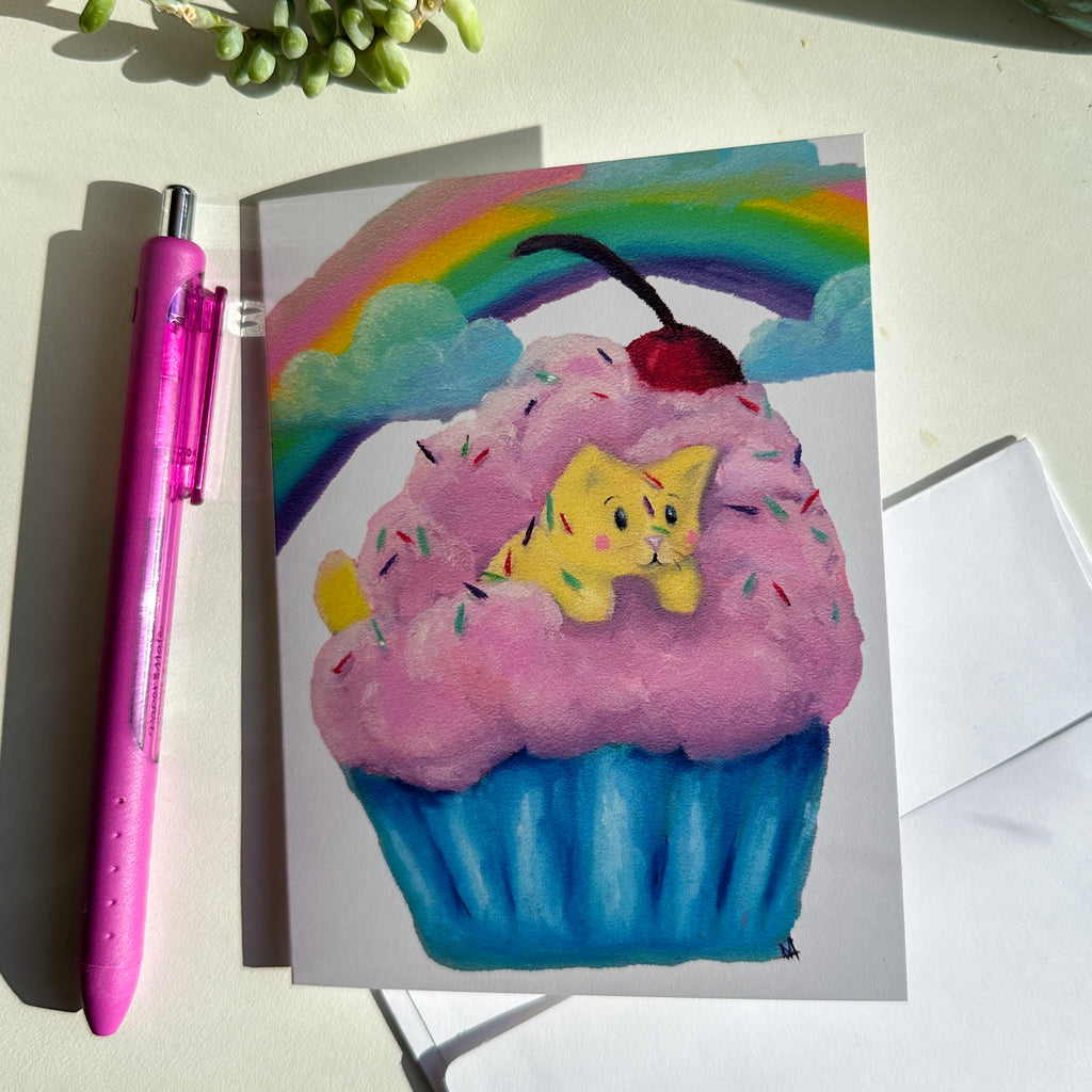 image of front of card with the yellow cat in a fluffy cupcake with pink icing and a blue cupcake paper.  There is a cherry on top along with sprinkles. There is a rainbow and clouds in the background. The card is next to a pen and has the envelope sitting under it. 