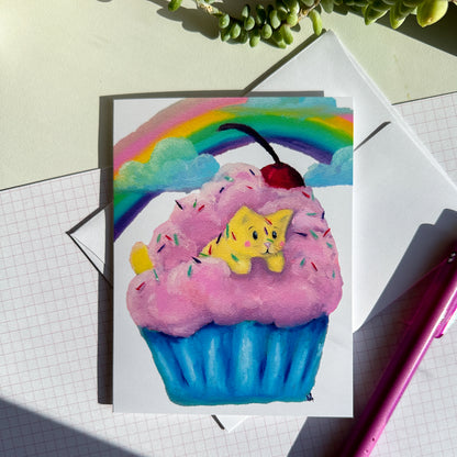 image of front of card with the yellow cat in a fluffy cupcake with pink icing and a blue cupcake paper.  There is a cherry on top along with sprinkles. There is a rainbow and clouds in the background. The card is next to a pen and has the envelope sitting under it. 